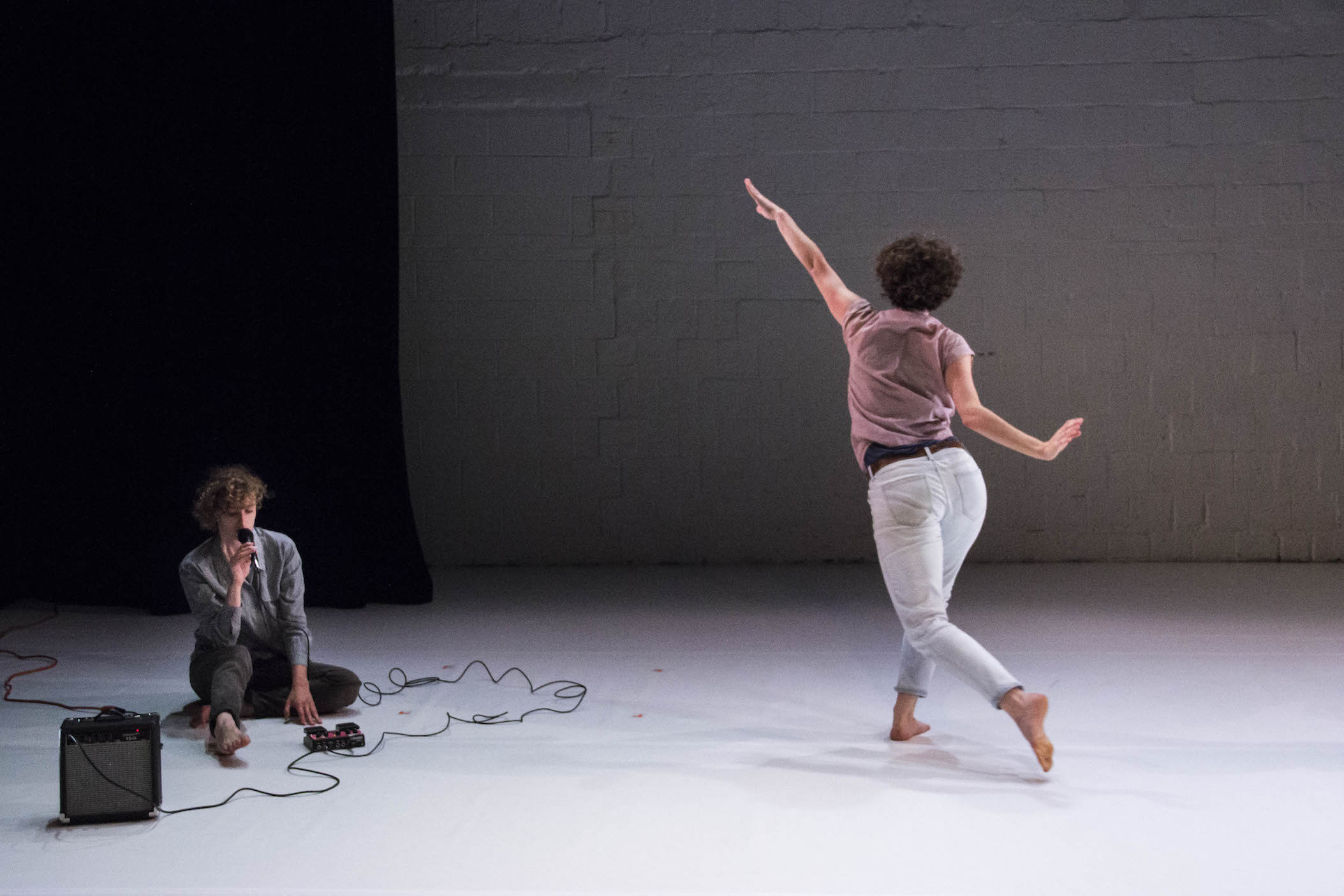 One dancer sits on the ground singing into a microphone, while another reaches towards an upstage white brick wall.