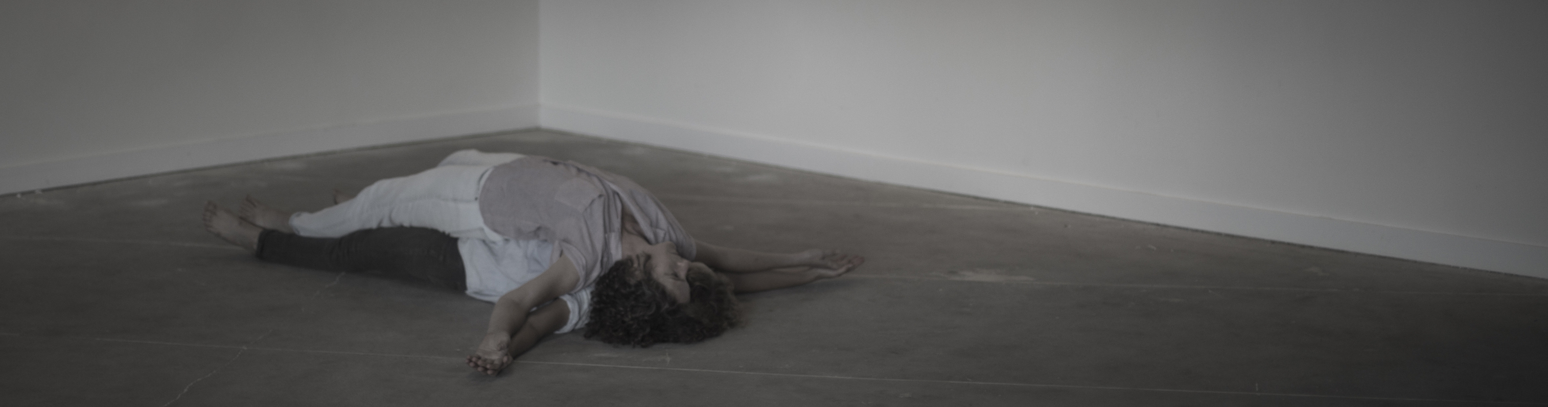 Two dancers lay stacked on top of each other on a grey concrete floor.