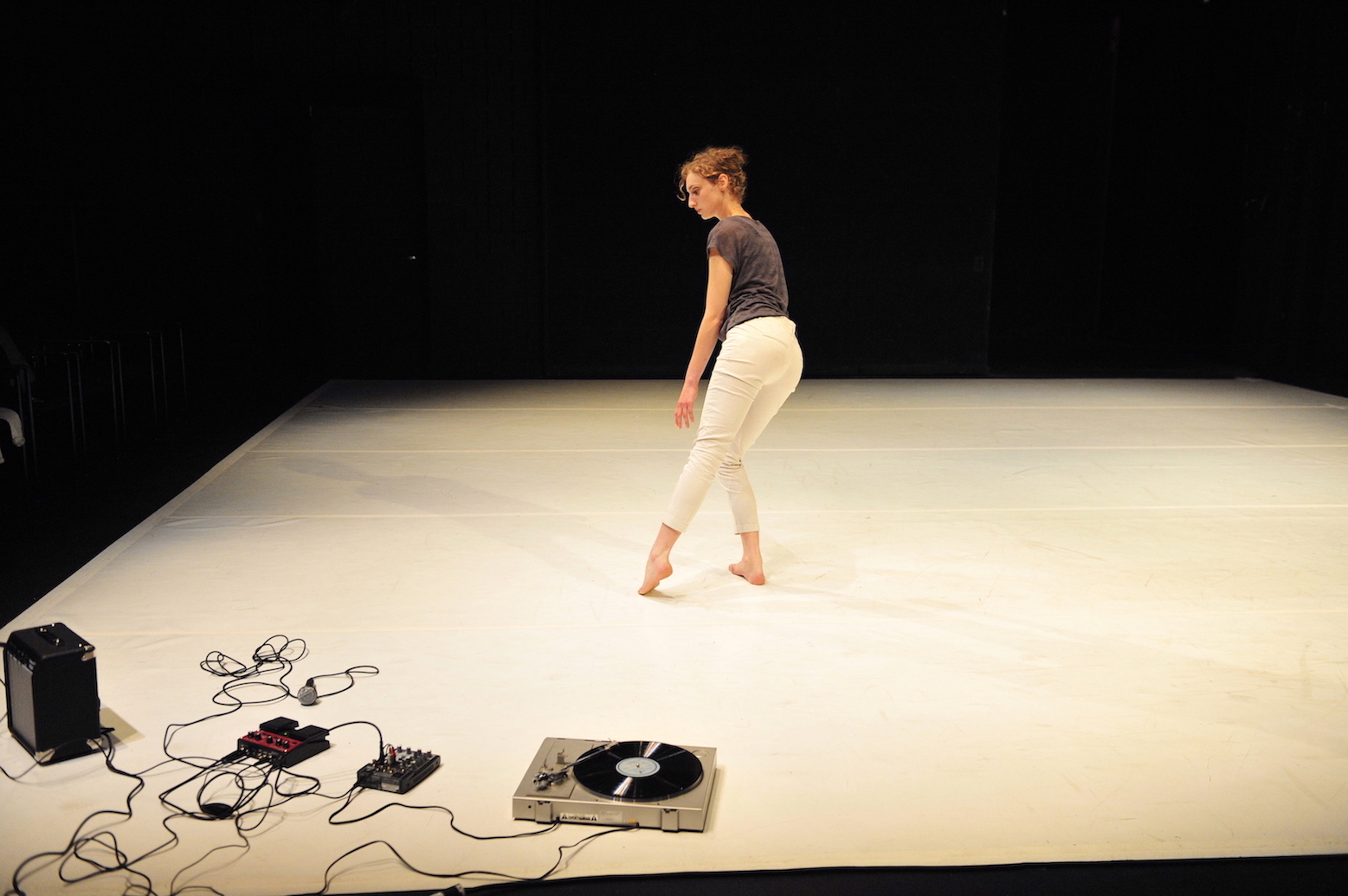 A white dancer wearing light pants and a gray tee shirt leans away from a turntable, microphone, looping pedal, mixing board, and cords in the corner of a white floor.