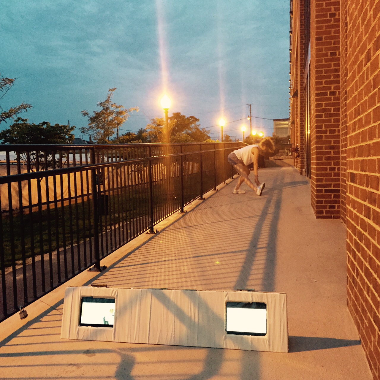 A white dancer lunges backward on a concrete train platform at dusk. In the foreground are two small video screens.