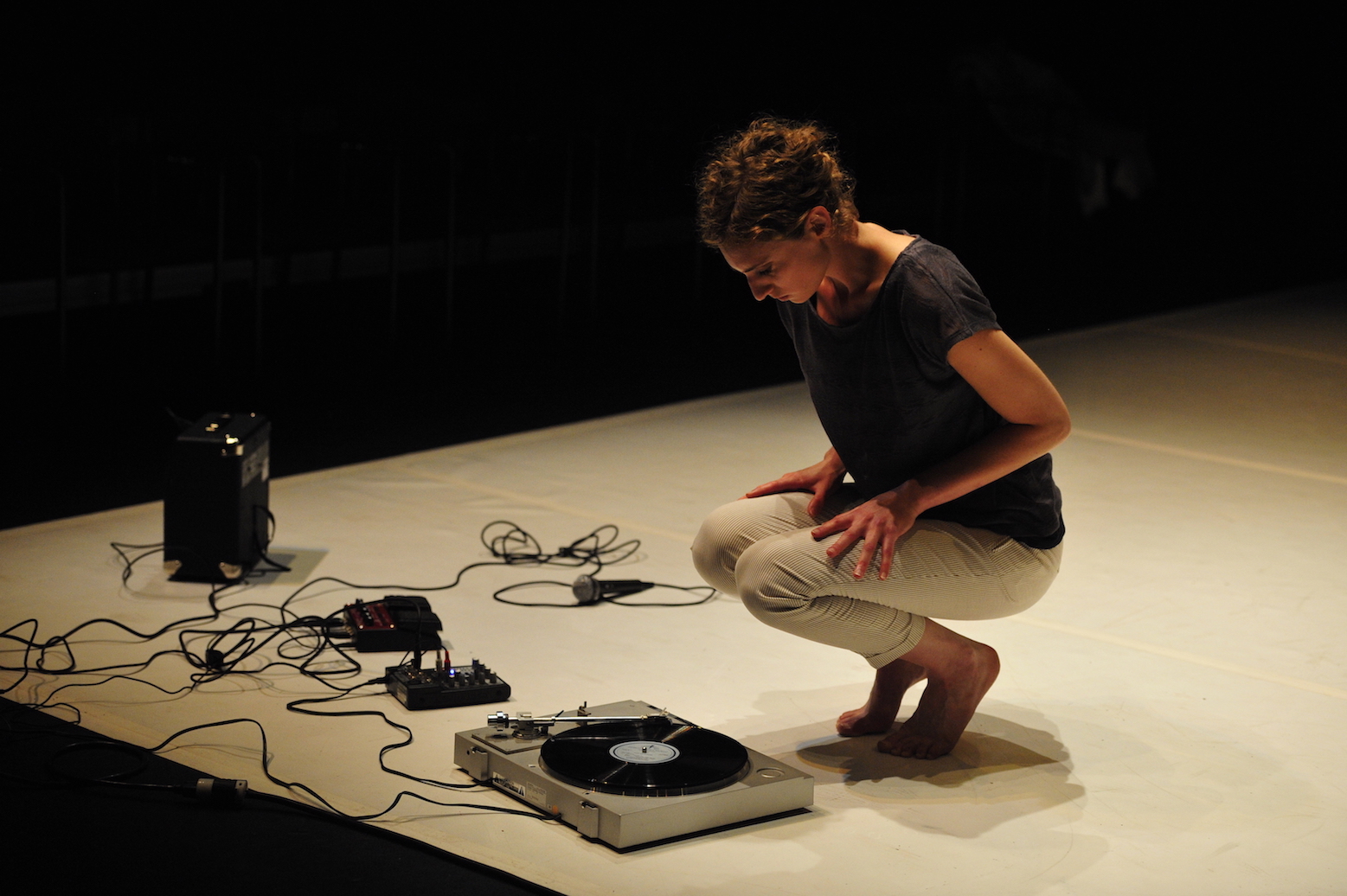 A white dancer wearing light pants and a gray tee shirt crouches over a turntable. A microphone, looping pedal, mixing board, and cords are spread out behind the turntable on white floor.