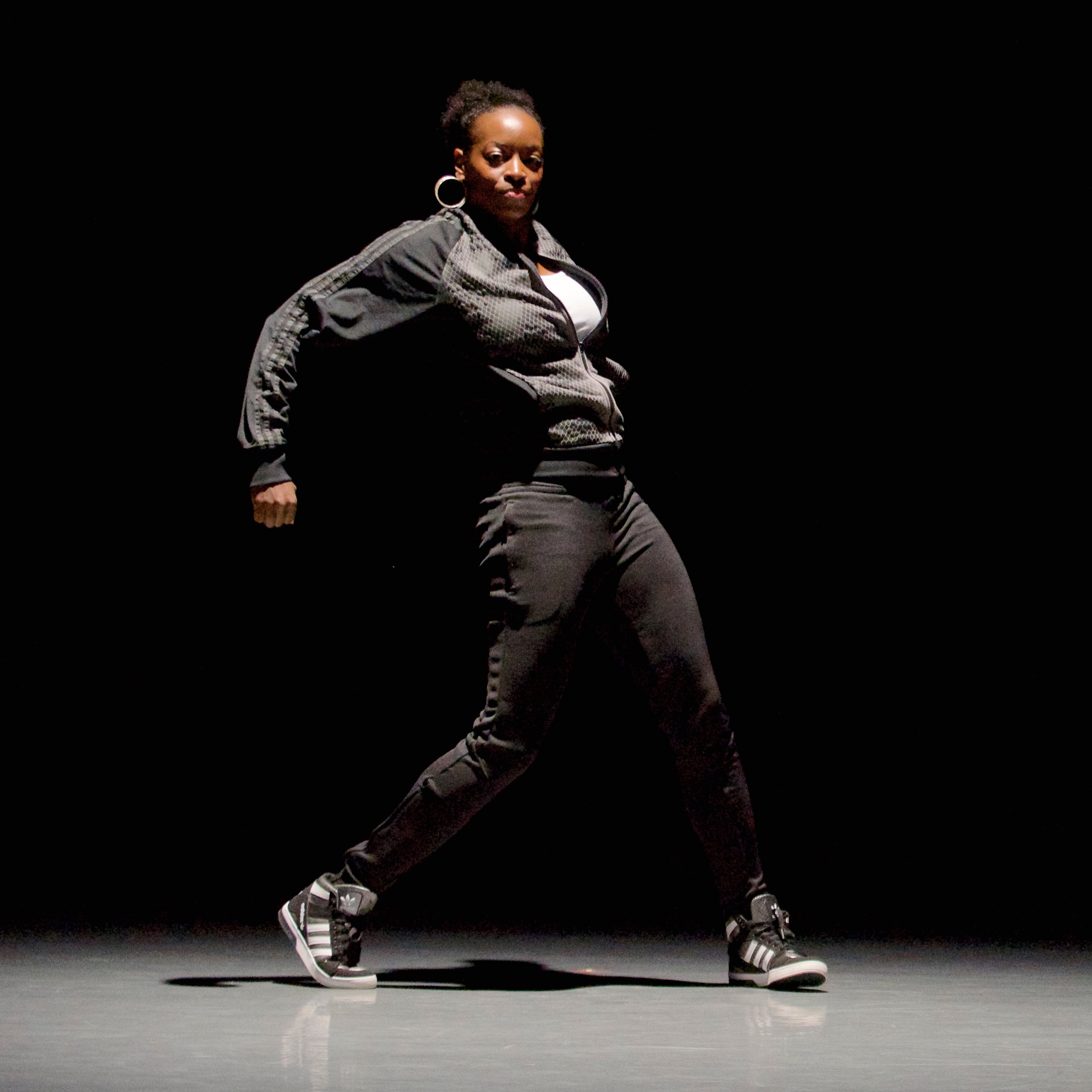 A woman dressed in dark gray/black poses with feet standing wide and arms shaped behind her