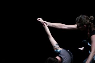 In a dimly lit image, one person reaches down to grasp the outstretched arm of another who is lying on the ground. The scene is dark, with only their arms and part of their bodies visible, creating a dramatic and intense atmosphere.