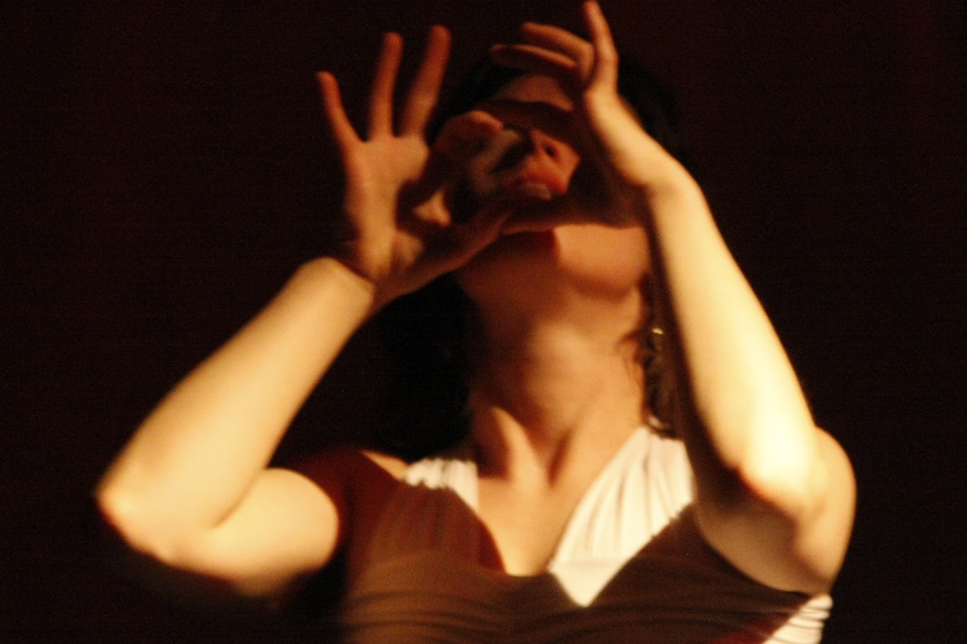 The image shows a woman in low lighting, her face partially obscured as she holds her hands near her eyes, forming circles with her fingers as if mimicking glasses. She is slightly out of focus, creating a mysterious and dramatic effect.