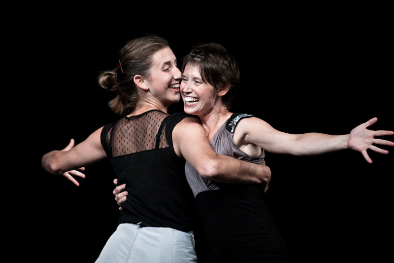 Two women joyfully embrace on a dark stage, both smiling widely. The woman on the left has her hair in a bun and wears a black top with sheer details. The woman on the right, with short hair, wears a sleeveless top. They appear happy and energetic.