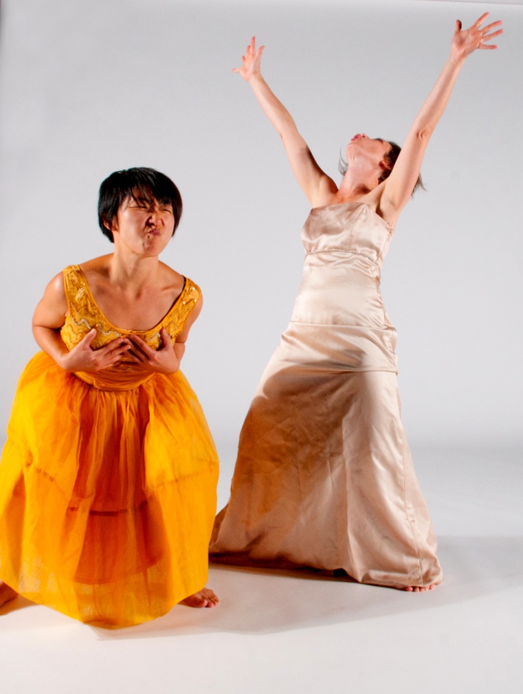 The image shows two women in expressive poses. The woman on the left, in a bright orange dress, makes a playful, exaggerated face. The woman on the right, in a beige dress, stretches her arms upward, looking towards the sky. The background is plain white.