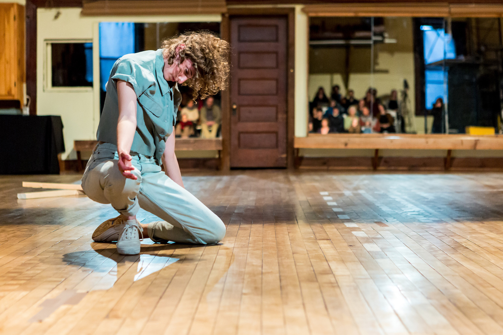 A dancer with curly hair dressed in light jeans, white sneakers, and a sage short-sleeved button down shirt kneels on the ground, arm outstretched and head tilted down, looking at hand. In the background, audience members are reflected in a mirror.