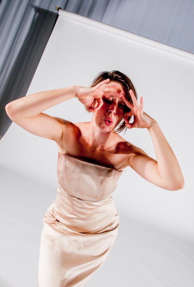 The image shows a woman in a strapless beige dress making a playful expression. She has short brown hair and holds her hands near her eyes, forming circles with her fingers as if mimicking glasses. The background is a white photo backdrop.