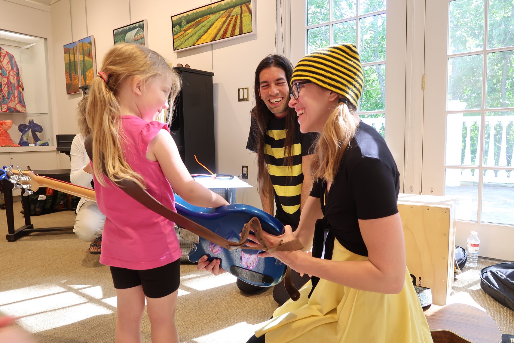 A musician dressed in bee colors shows a young child how to play a blue bass guitar