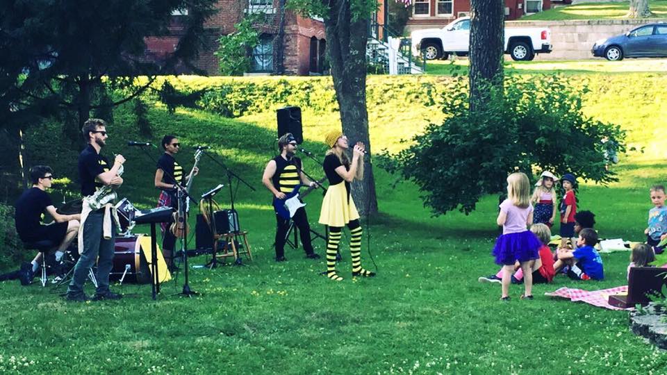 Musicians dressed in bee colors play on a park lawn with an audience of children in front of them