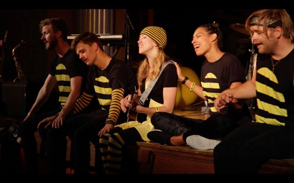 Musicians dressed in bee colors sit at the front of a stage with hands on each other's shoulders