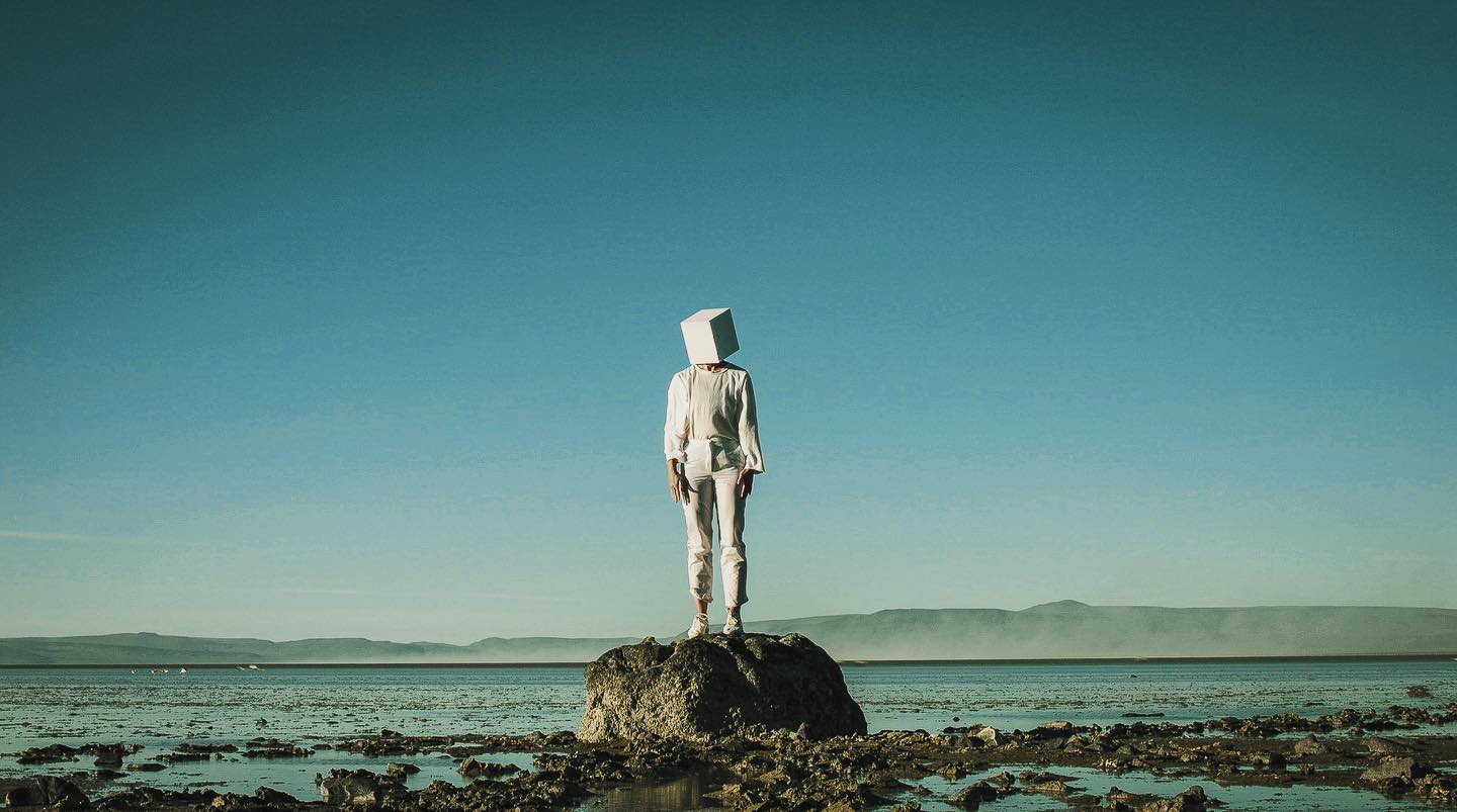 dancer Ellie stands on top of a rock with a blue sky behind her, she wears an all white outfit with a white cube on her head