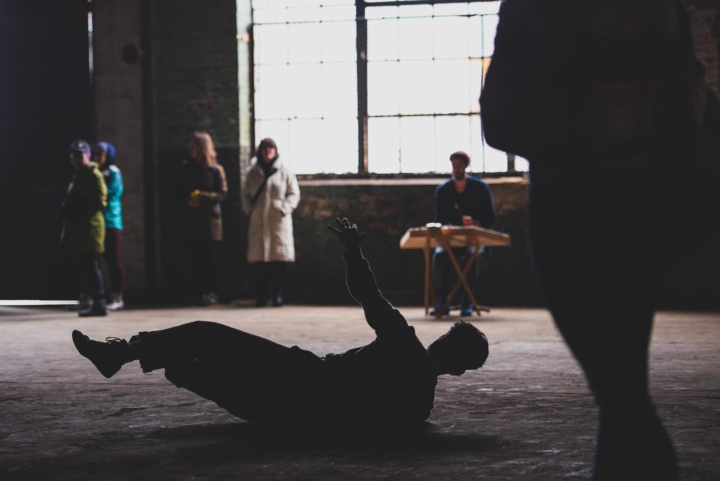 dancer Jungwoong Kim rolls on the floor during a performance of "JUNKSPACE" in Philadelphia while musician Cole, behind Jungwoong, plays the hammered dulcimer and audience members watch