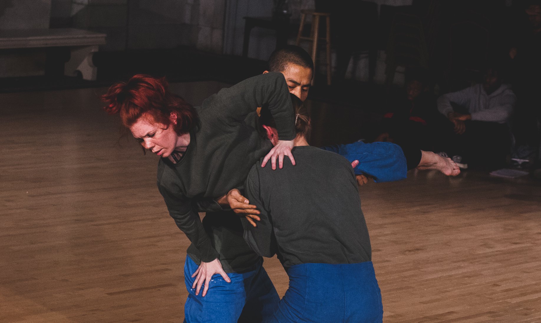 Two dancers lift one dancer between them, everyone wears blue pants and a dark green top
