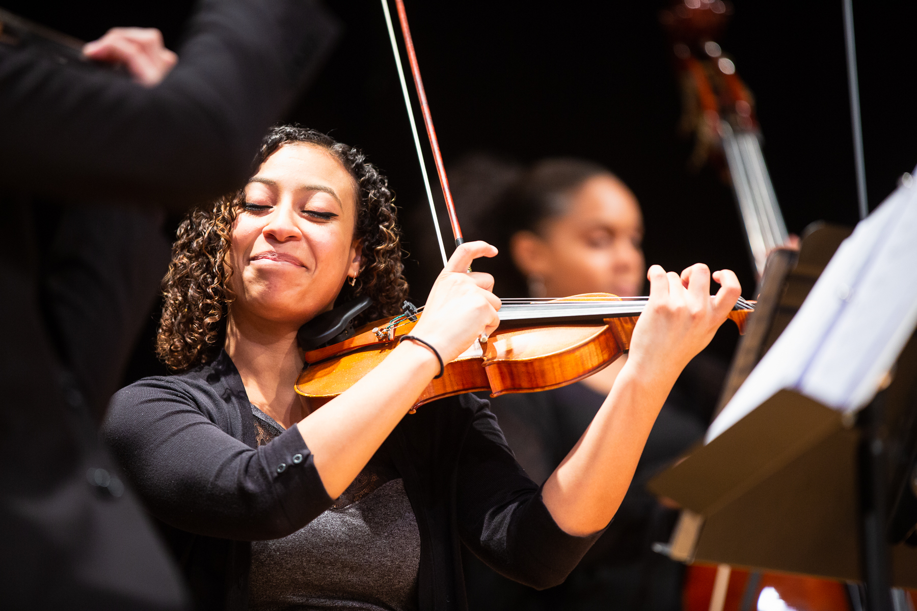 A Black female with curly brown hair holding a violin. Her eyes are closed and she is smiling while facing the camera.