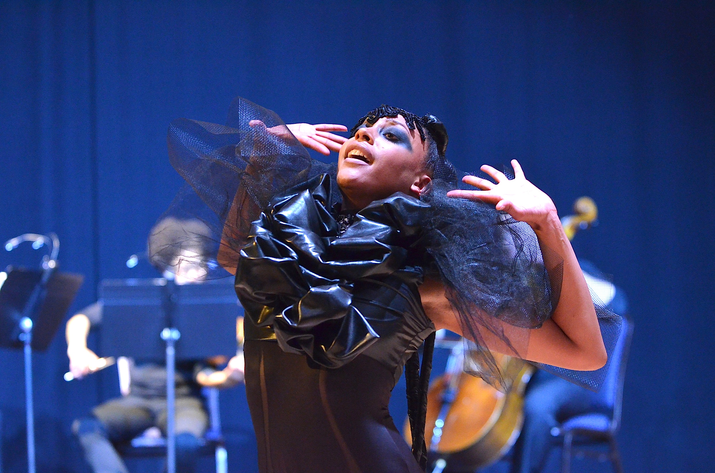 A Black female dancer with arms stretched and chest puffed out wearing a black, decorative costume. A group of string players perform behind her.