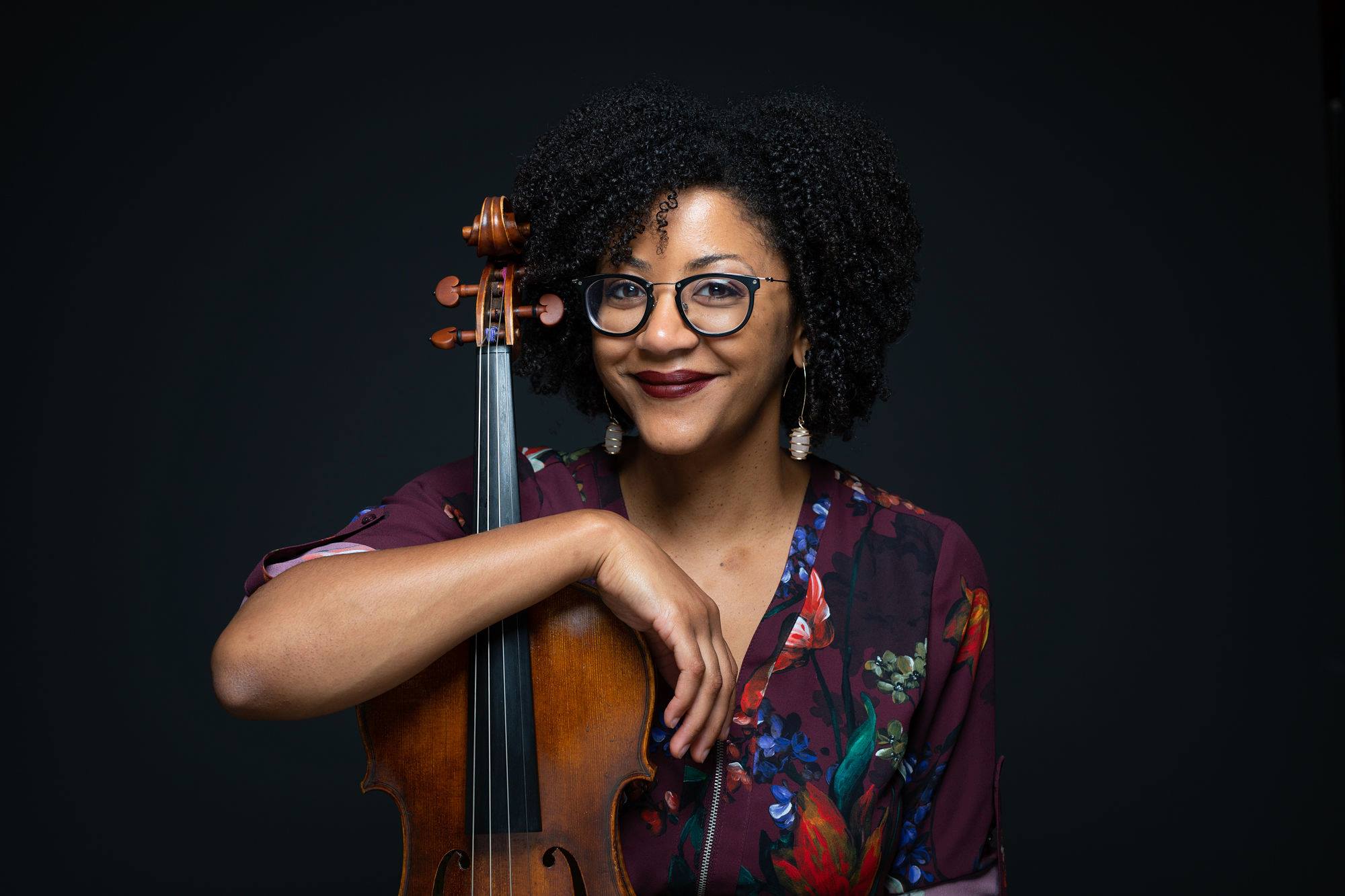 A Black woman with curly hair wearing a flower dress holding a viola. Caption: Ashleigh Gordon, Artistic Director & Violist