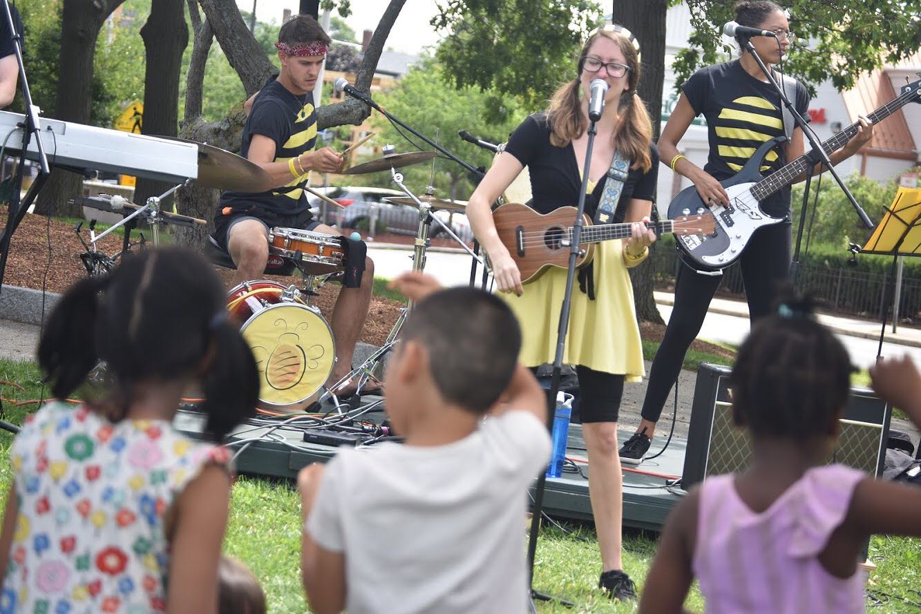 Image shows a singer dressed in black and yellow playing a ukulele outdoors, with band members behind her and kids dancing in the foreground