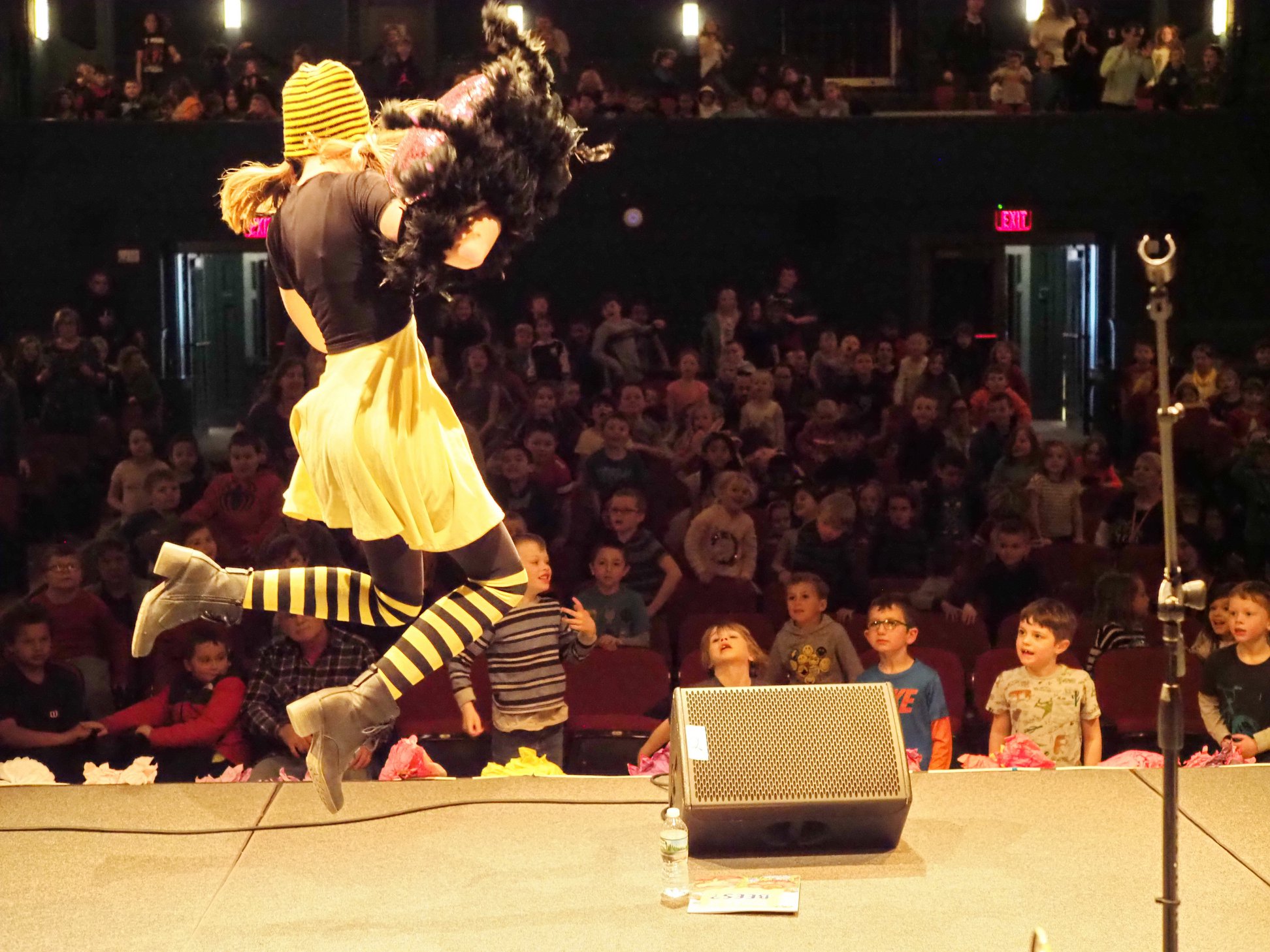 Musician dressed in bee colors and holding a spider puppet jumps in the air in front of a crowd of children