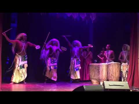 Rwandan men dance in long skirts with three-foot long grass head dresses as drummers and female dancers play music.