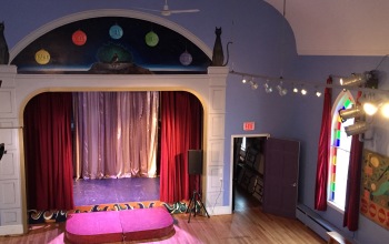 Mayo Street Arts' proscenium stage can be seen, adorned by a trompe l'oeil mural depicting the moons of pluto, which was painted on MSA's periwinkle walls by Pat Corrigan.