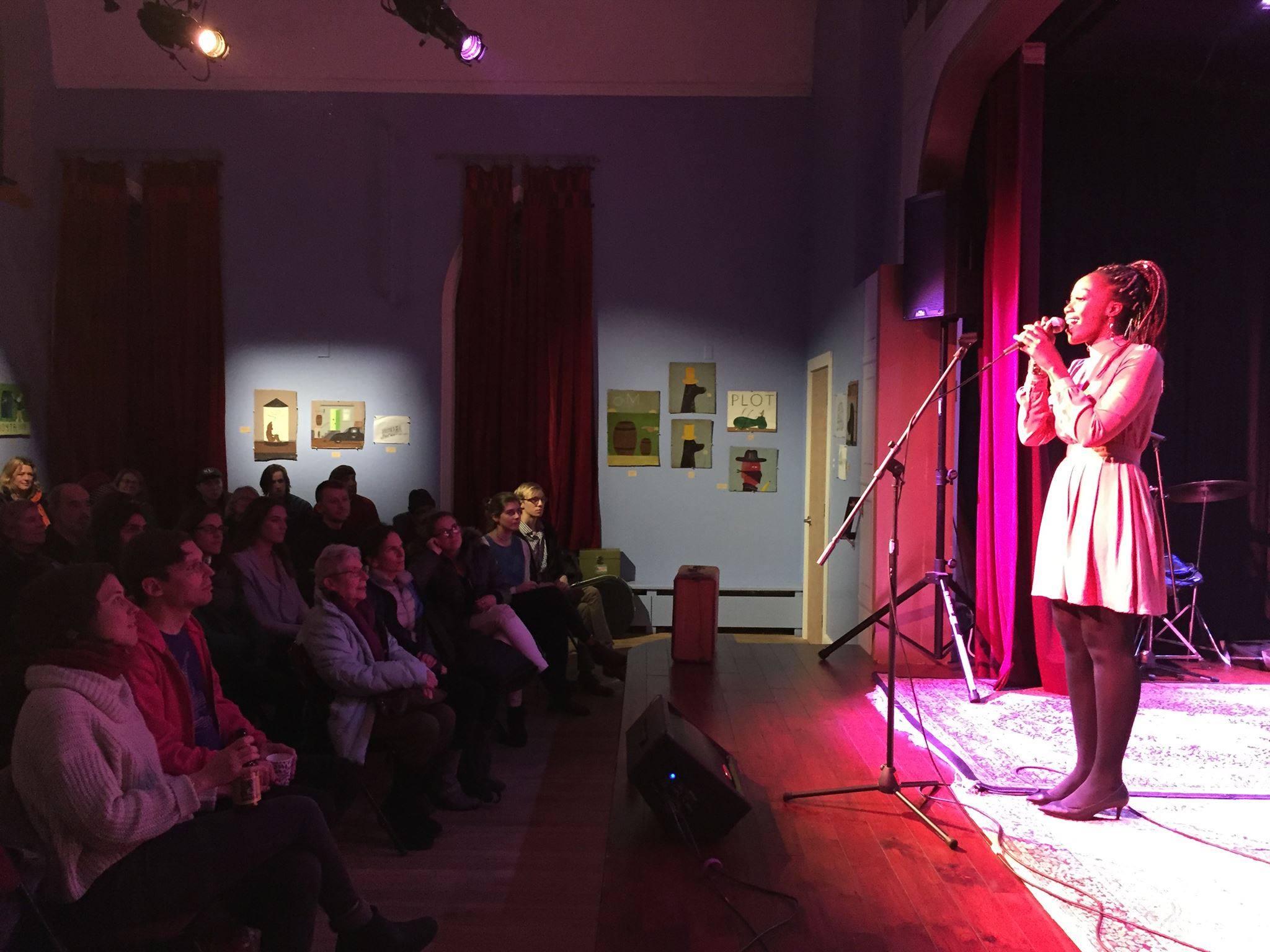 A woman of color holds a microphone close to her face as she sings for an full audience at Mayo Street Arts.