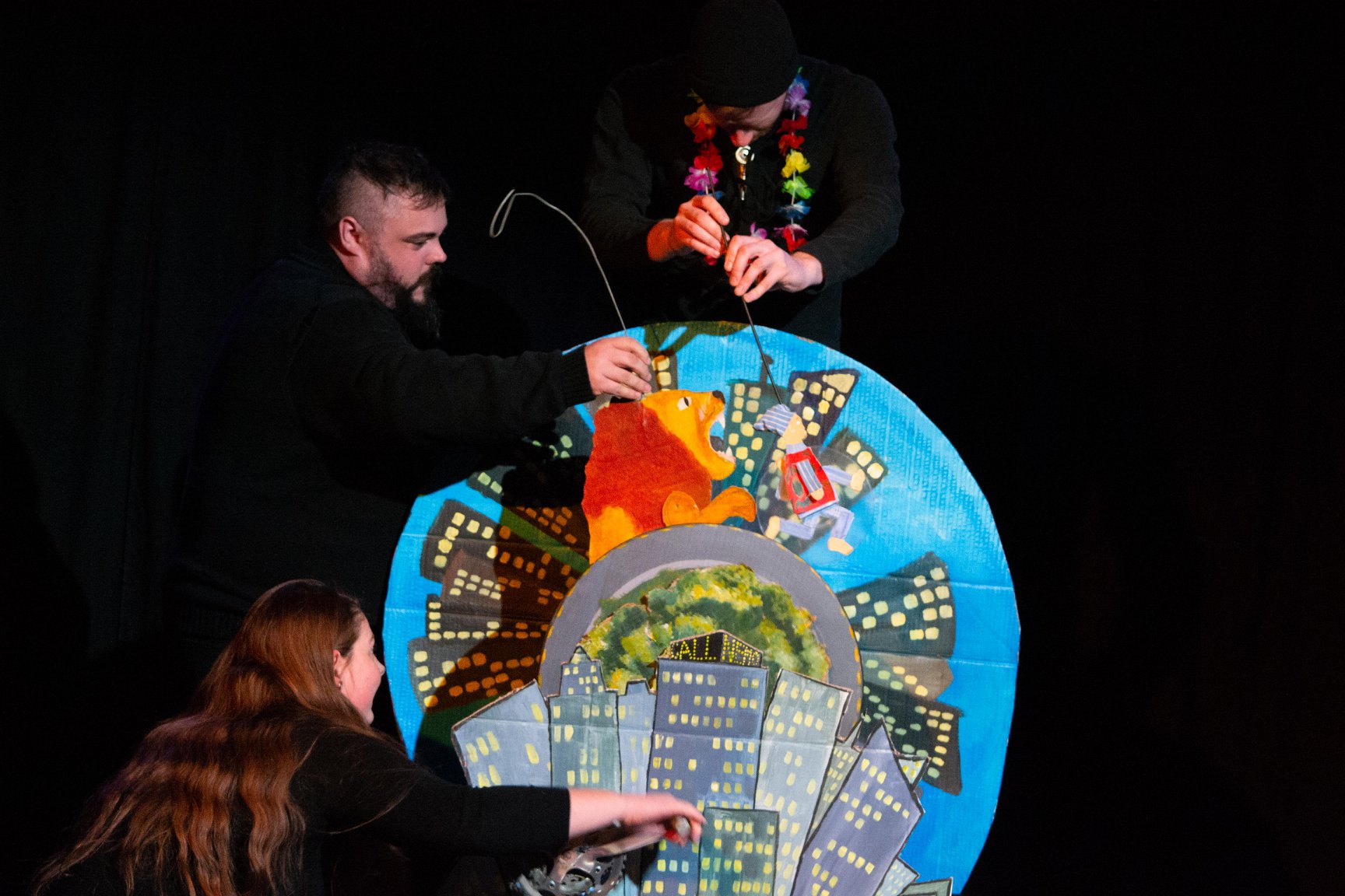 Three puppeteers dressed in black clothes animate a lion chasing a man in a stocking cap who run in front of an endlessly spinning circular set.