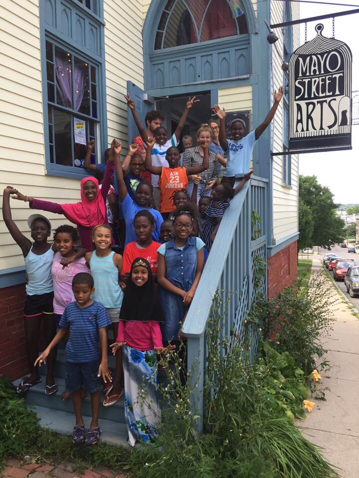 Students wearing summer clothes, some of whom sport head coverings, raise their arms in joy on Mayo Street Arts' front steps.