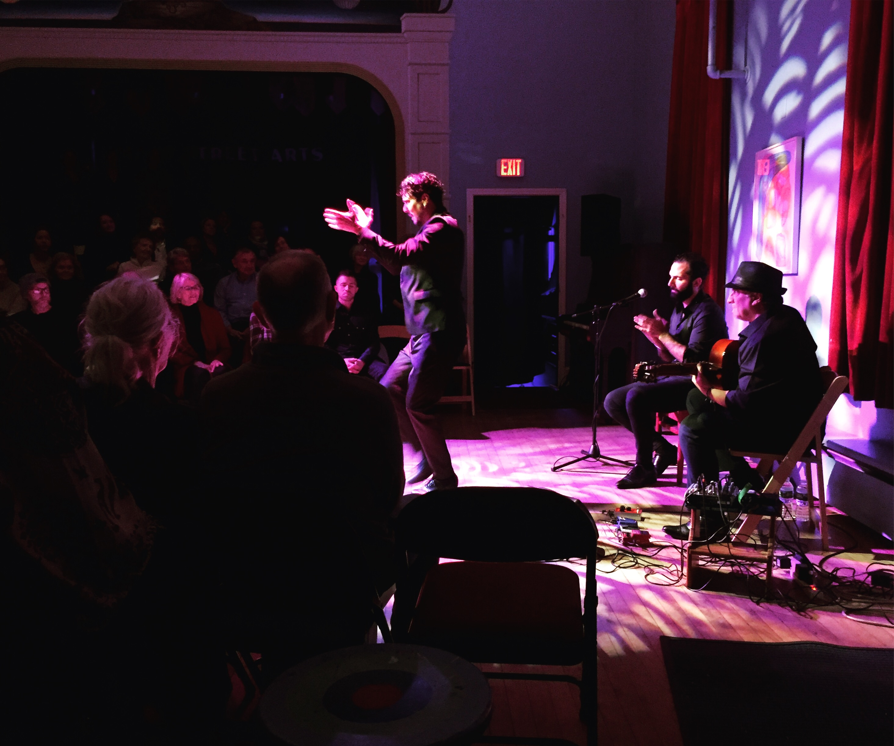 A male Flamenco dancer stomps and claps in a purple light, surrounded by the audience and a pair of seated musicians.