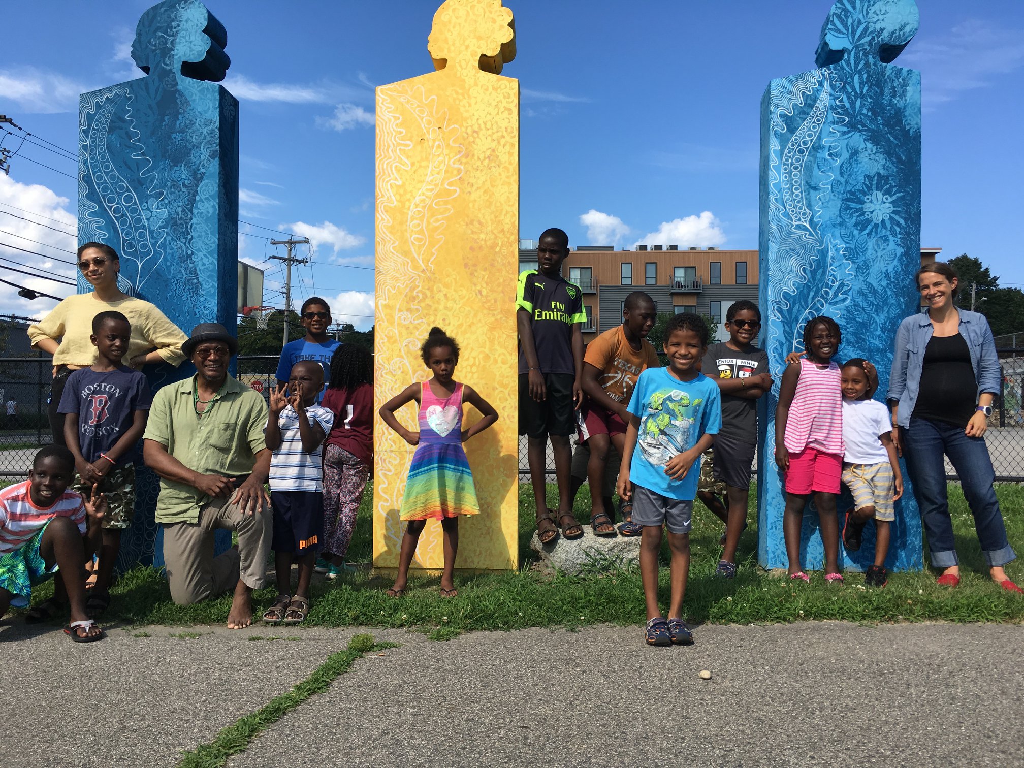 Children in colorful, summer clothing pose in front of large, sculptures with the artist. The sculptures are monoliths with BIPOC bust silhouettes at the top.
