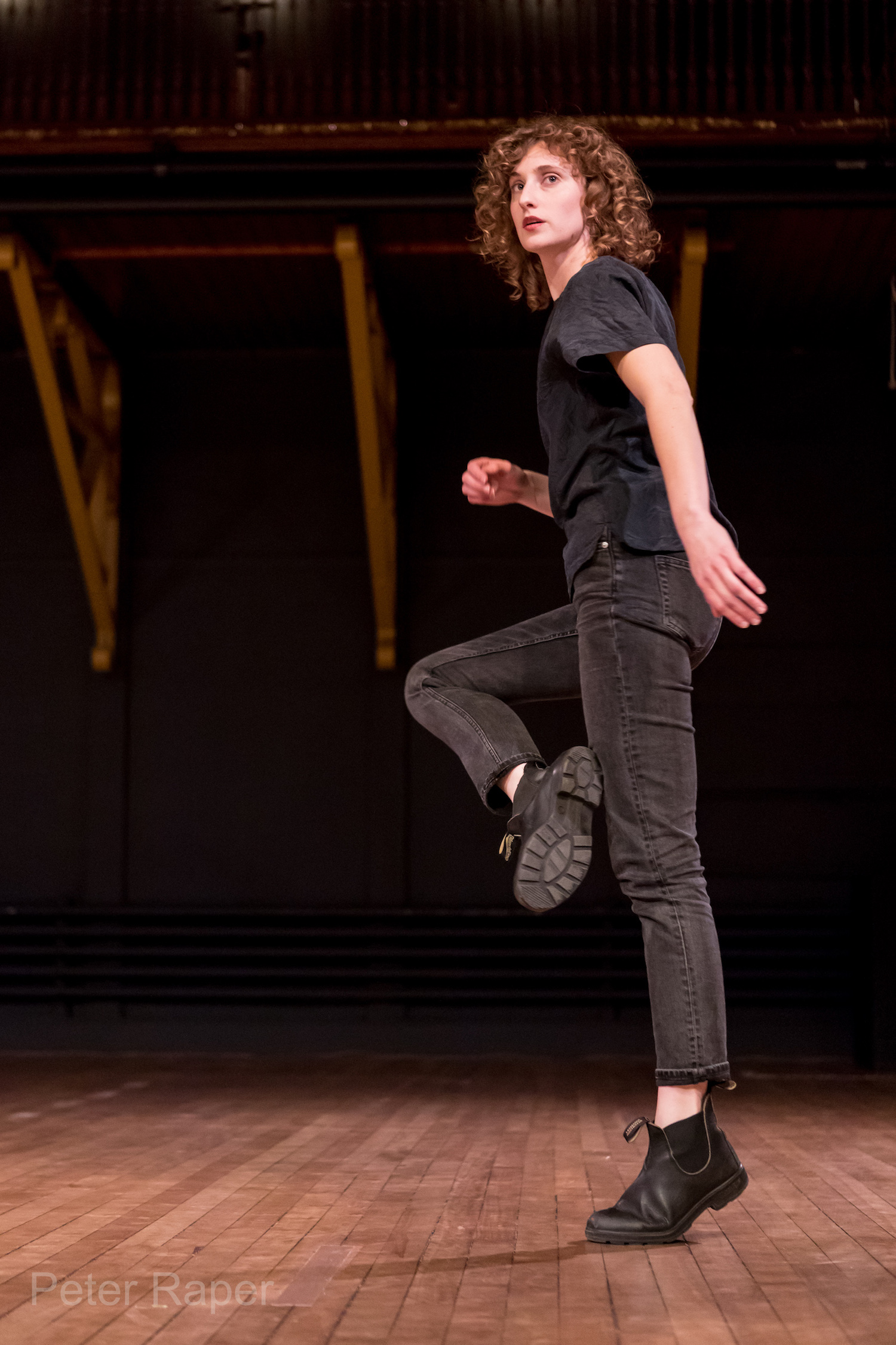 A white dancer dressed in black jeans, black tee shirt, and black boots with mid length light brown curly hair stands on one leg while leaning away, uplifted knee bent. She looks back toward the audience.
