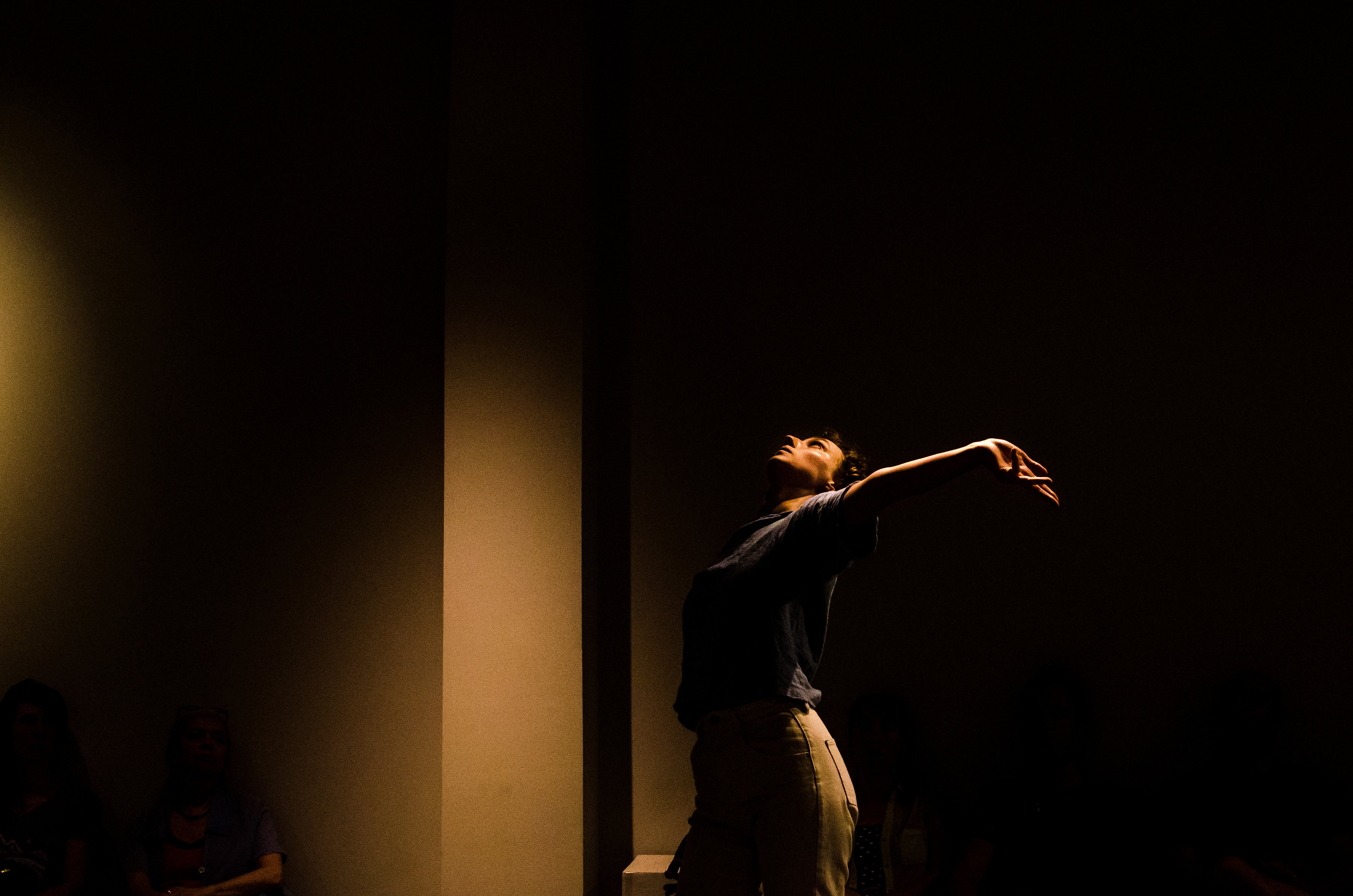 A white dancer in a dimly lit space reaches out her left arm with hand and fingers flexed back while looking up at the ceiling.