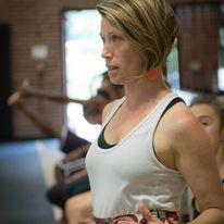 A woman with short, highlighted hair, focused and standing in a bright, indoor setting. She wears a white tank top with a black sports bra underneath. Other blurred figures are visible in the background, suggesting a group setting or class.