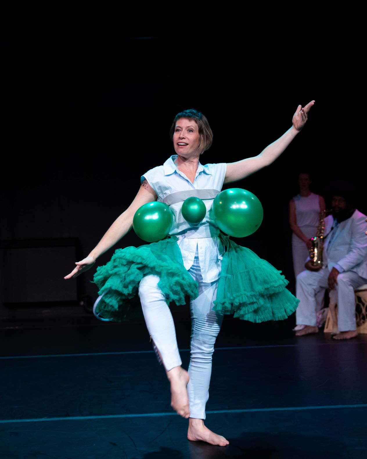 A woman performs on stage in a white outfit with green balloons attached to her torso and a green tutu. She stands on one leg with one arm extended, looking playful. The background is dark, with two other people faintly visible.