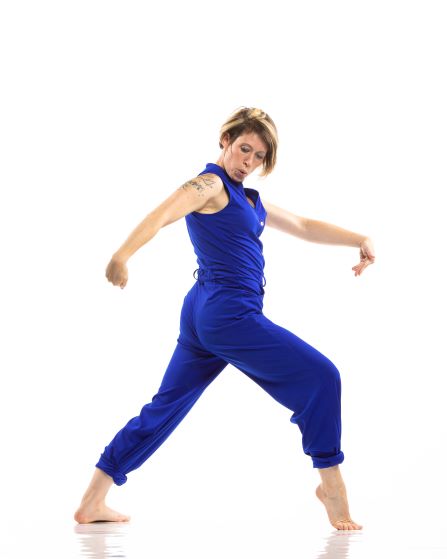 The image shows a woman in mid-dance, wearing a vibrant blue jumpsuit. She has short, highlighted hair and a tattoo on her upper left arm. Her posture is dynamic, with one leg extended back and arms gracefully positioned. The background is plain white.