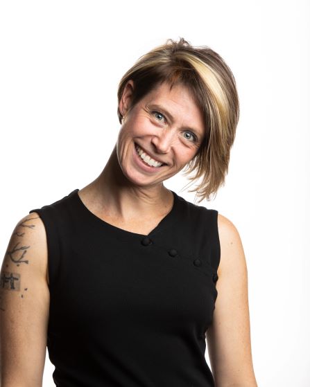 The image shows a smiling woman with short, light brown hair with blonde highlights, wearing a sleeveless black top. She has a tattoo on her upper left arm. The background is plain white, and she appears cheerful and approachable.