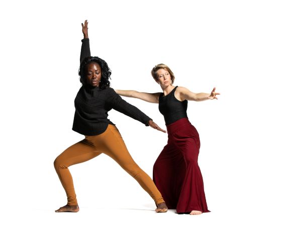 The image shows two women striking dynamic dance poses. The woman on the left, with long dark hair, wears a black top and orange pants. The woman on the right, with short hair, wears a black top and red flowing pants. Both appear focused and expressive.
