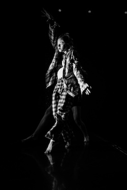 White cis-woman dancer wearing plaid pants and a plaid shirt unbuttoned with a white tank top closes her eyes, reaches up her right arm, opens her left palm, and steps forward on her left toes.  She is sensing the space in front of her.