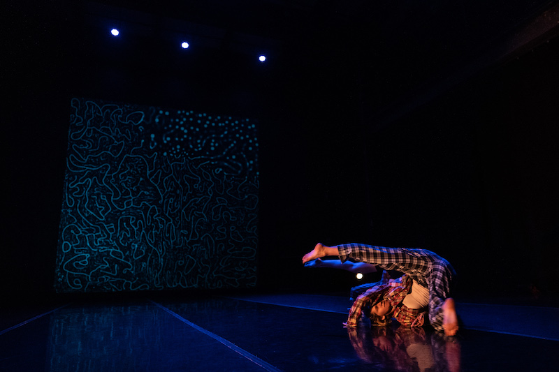White cis-woman dancer rolls over her shoulder and kicks her leg in the air in low light.