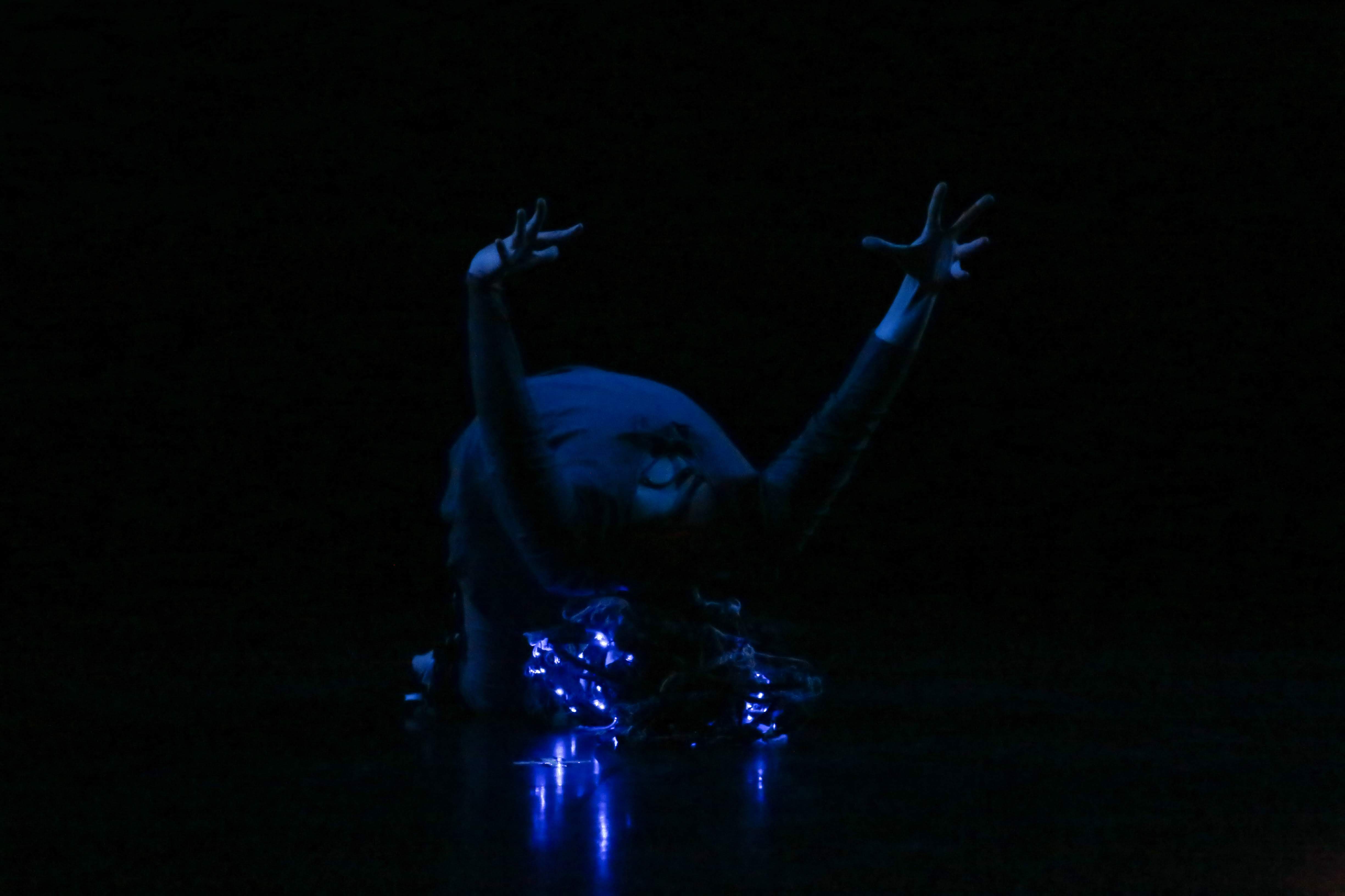 Dancer on the floor in the dark, bowed forward with arms extended behind her, wearing a light up headpiece
