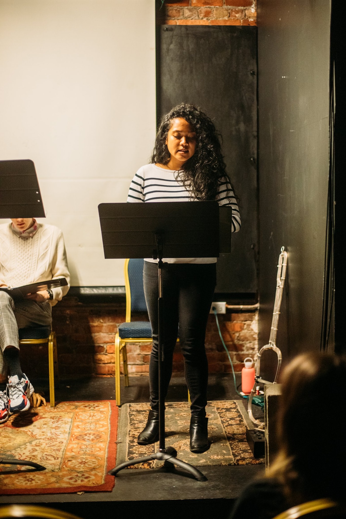 Aurora on stage, standing and reading from a stand. She’s dressed in a striped black and white long sleeve top and black jeans, with curly hair resting on her left shoulder, under stage lighting.