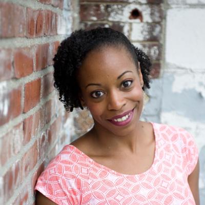 A woman (Shakia Barron) leans against a brick wall, dressed in pink and pictured from the shoulders up