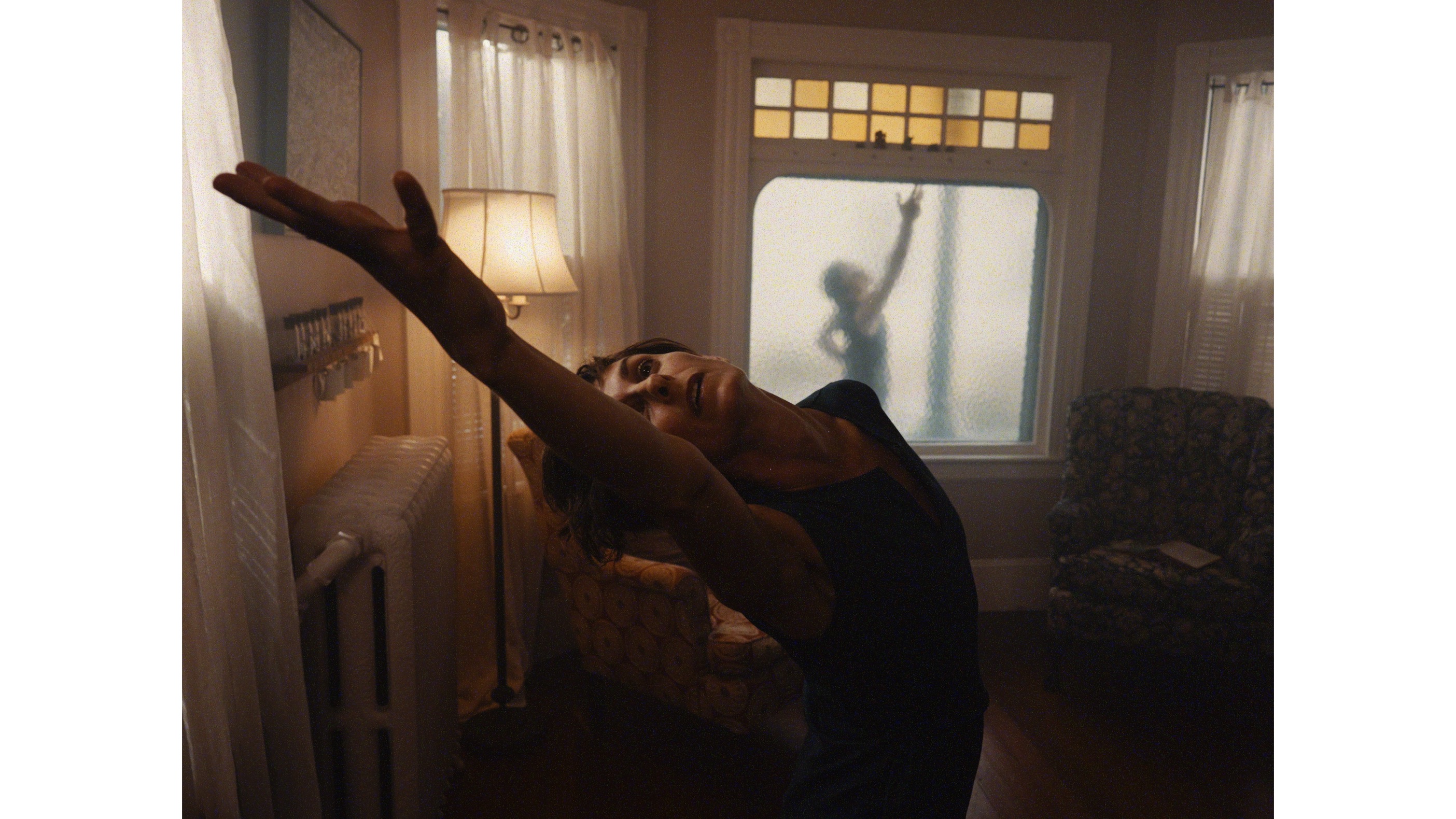there is one woman visible in the foreground with arm outstretched contrasted with a woman in the background seen behind a plane of glass whose arms is also outstretched.. 