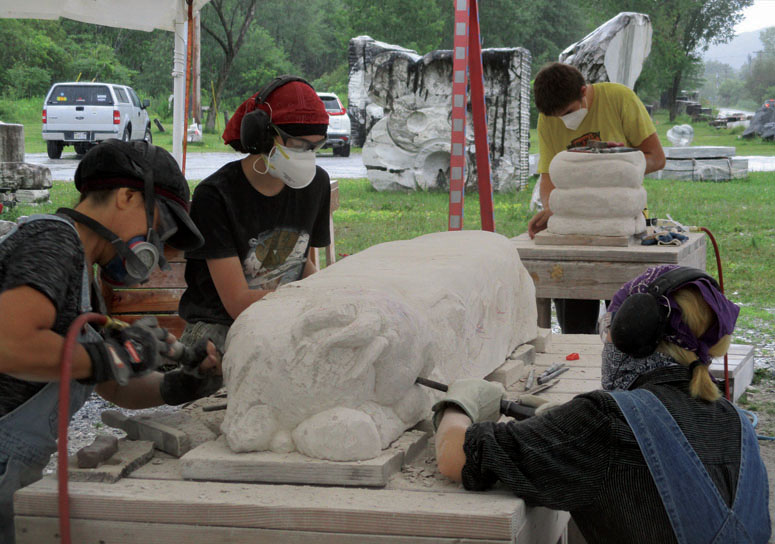 a group of teens work outdoors with power tools to carve a bench from granite
