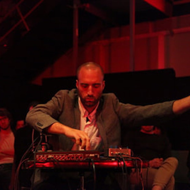 A white man in a grey suit jacket and white shirt performs, manipulating a mixing board with cords coming out the back of it. His left arm reaches to the side. There is reddish light filling the space. 
