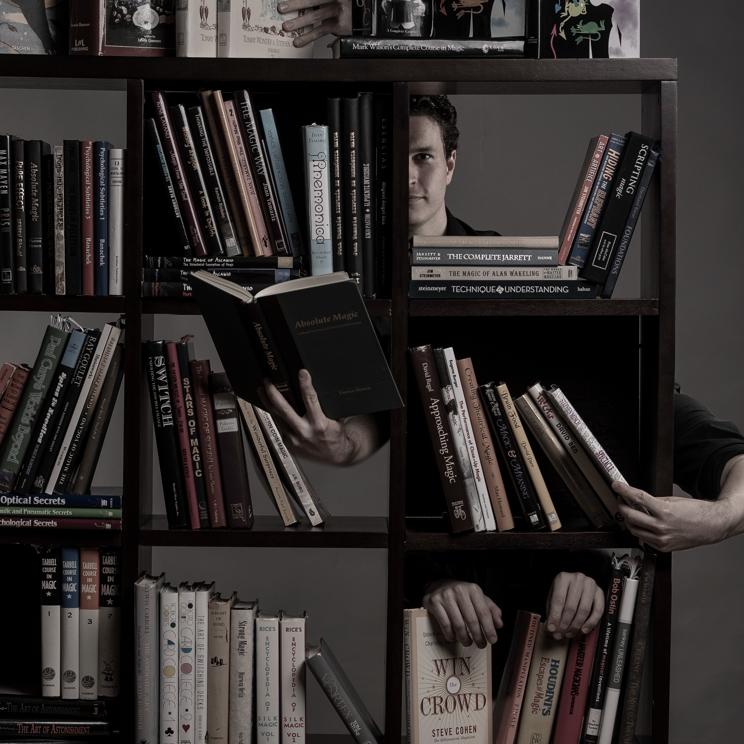 A bookshelf full of magic books in the foreground. Evan's face peeks through the back and an impossible number of hands reach around to different books.