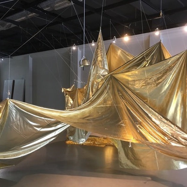 Shiny, golden fabric fills a gallery with a grey floor and white walls. The fabric is suspended from the ceiling, creating triangular peaks and rippled slopes.