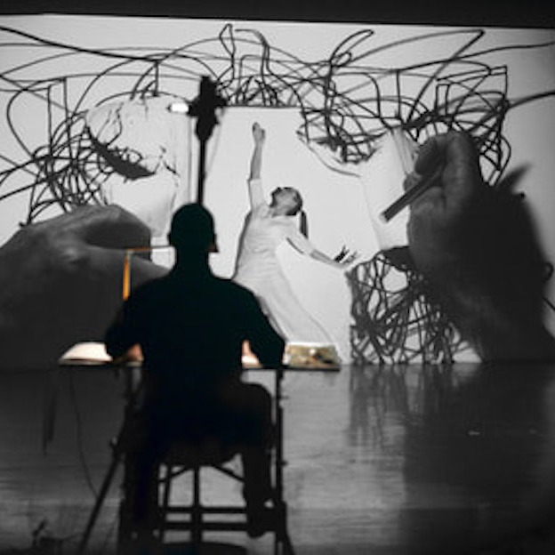 A black and white image with a seated man silhouetted. He faces a dancer in a white dress. Her arms are extended and she looks up. Behind her, there is video projection showing hands scrawling looped, intersecting black lines on a white background.  
