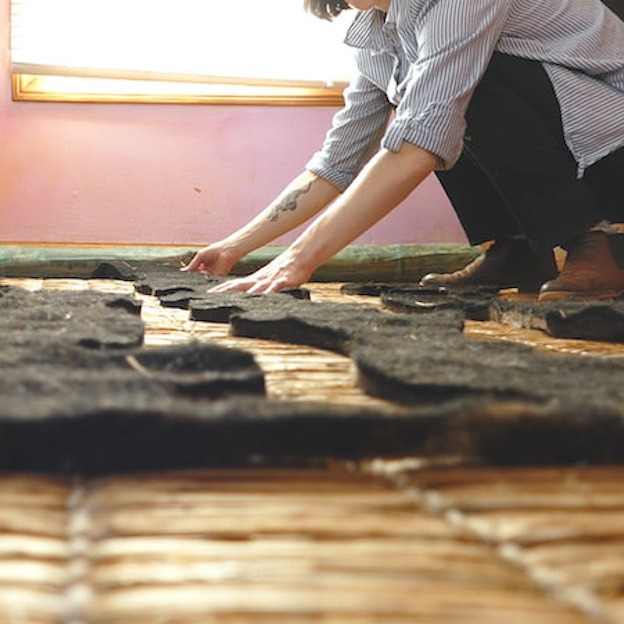 Cut felt shapes lie on a brown woven mat on the floor. A white woman's arms are visible holding one of the shapes. She wears a light blue shirt and has a tattoo on her right forearm. 