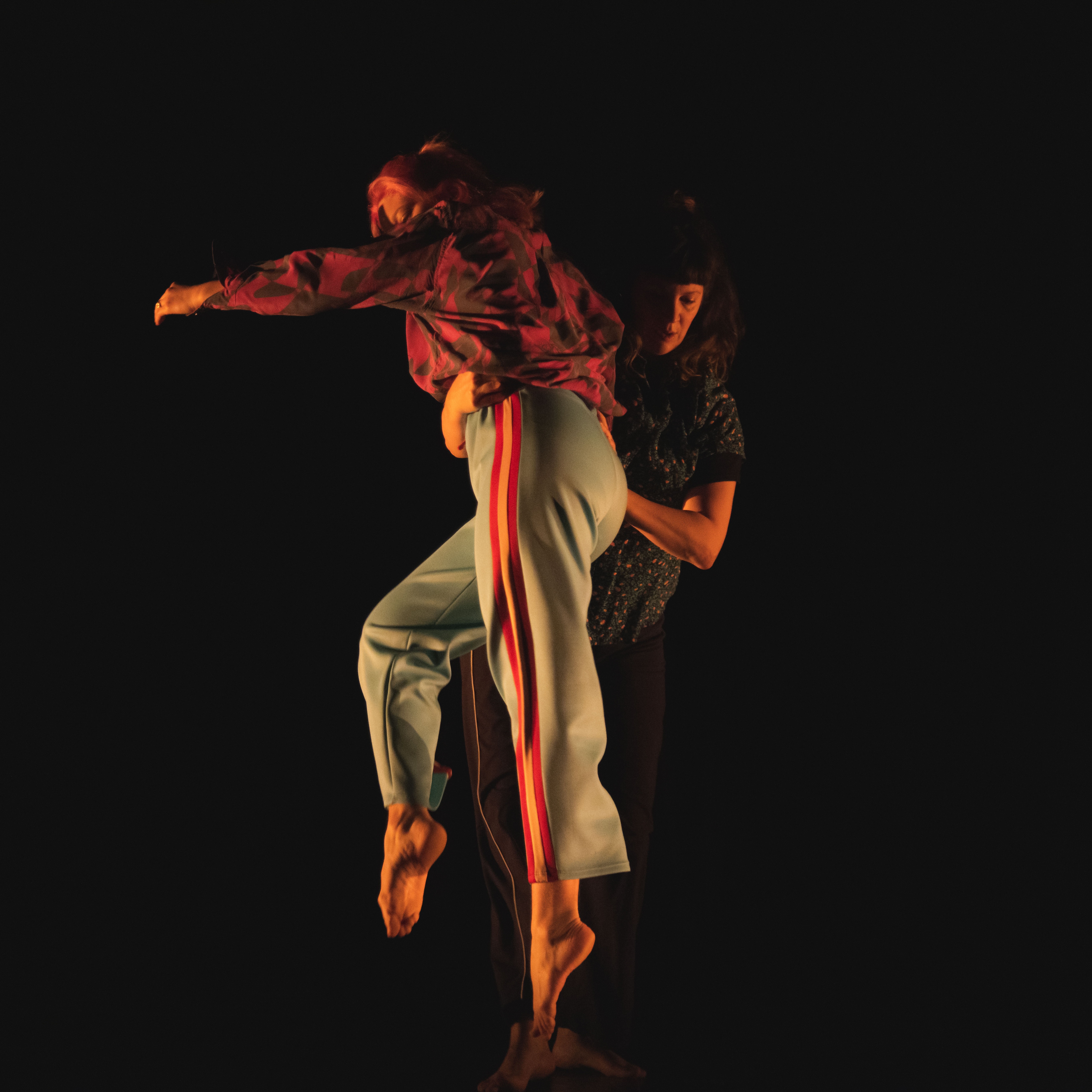 2 white cis-women wearing floral shirts and track pants are dancing contact improvisation. 